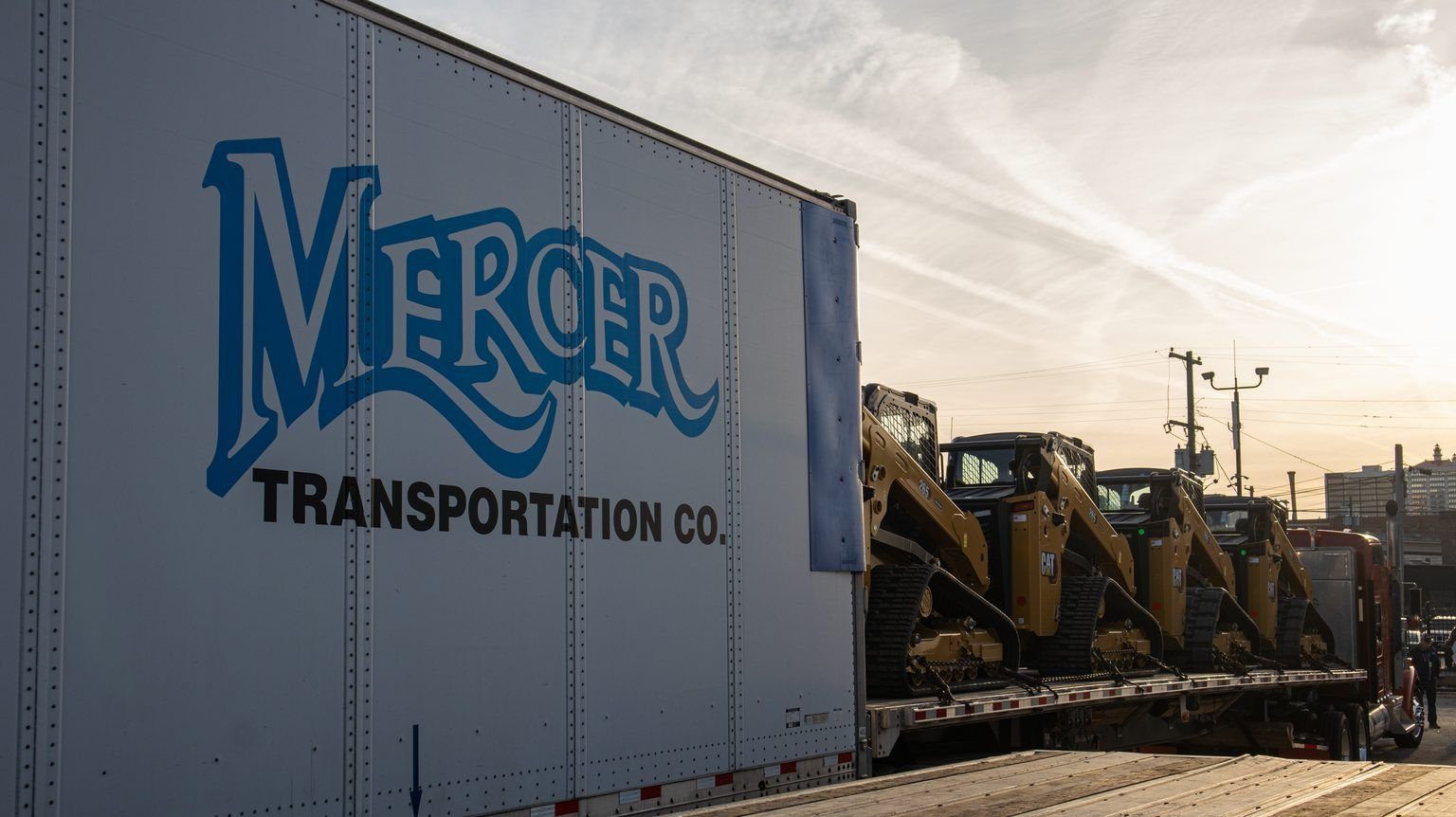 Caterpillar skid steers secured on a Mercer flatbed beside a Mercer Transportation van trailer